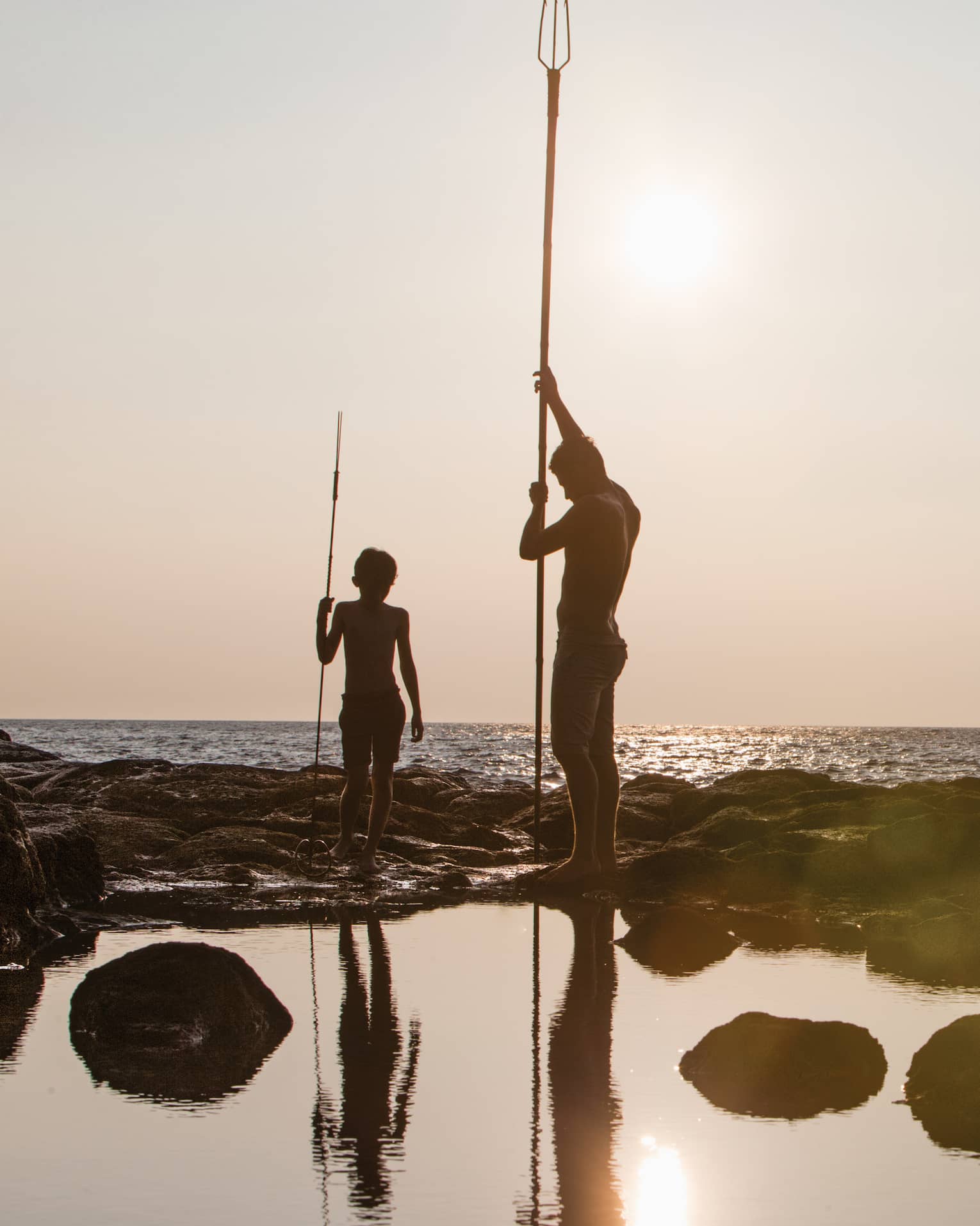 Silhouettes of people holding long poles on rocks near ocean at sunset